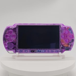 Pode incluir: Uma consola de jogos portátil com uma estrutura de plástico roxo translúcido. O dispositivo apresenta um ecrã preto central, um pad direcional à esquerda, quatro botões de ação à direita e os logótipos da Sony e PSP na parte frontal.