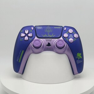 Pode incluir: Um comando de videojogos sem fios com um design em roxo escuro e lavanda claro. O painel tátil central apresenta o número 001 e texto gráfico. O lado direito apresenta um logótipo NERV verde, enquanto o lado esquerdo tem texto verde.