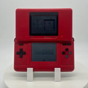 May include: A red Nintendo DS handheld game console, open to display two screens. The top screen shows a clock and calendar, while the bottom screen displays a menu. The console is supported by a white stand.