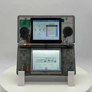 Puede incluir: Una consola de juegos portátil gris transparente con dos pantallas, que muestra un reloj y un calendario en la pantalla superior y un menú en la pantalla inferior. La consola está abierta, revelando componentes internos y altavoces. El dispositivo está sobre un soporte blanco.