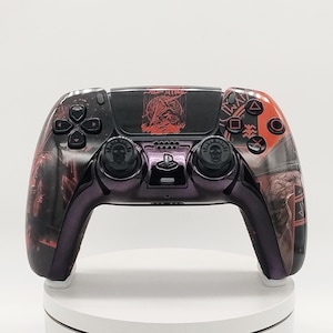 May include: A wireless video game controller featuring a custom black and red graphic design. The central touchpad displays a red illustration of a hooded figure. The analog sticks are black with skull icons, and the controller body has a metallic purple finish.