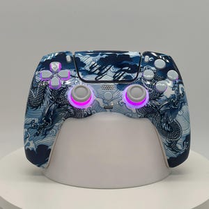 May include: A custom video game controller with a blue and white dragon design. The controller features a detailed dragon and wave pattern, with illuminated buttons and thumbsticks. The controller is resting on a white stand.
