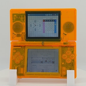 Pode incluir: Console de jogos portátil Nintendo DS laranja, aberto para exibir duas telas. A tela superior mostra um relógio e um calendário, enquanto a tela inferior exibe um menu. O console tem uma carcaça de plástico transparente.