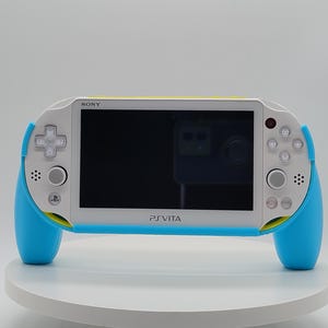 Può includere: Console di gioco portatile PlayStation Vita bianca e blu. La console ha uno schermo nero, un pad direzionale e diversi pulsanti. Le impugnature sono blu brillante e il bordo superiore è giallo. La console è su una superficie bianca.