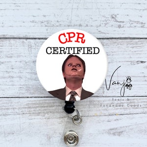 May include: A white button with a red "CPR Certified" text and a picture of a man with a surprised expression. The button is attached to a black retractable badge holder.