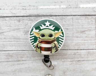 May include: A round badge reel featuring a green and white Starbucks logo with a cartoon image of Grogu, also known as Baby Yoda, holding a coffee cup. The badge reel has a black retractable cord and a clear plastic strap with a metal snap.