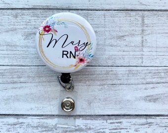 May include: A white retractable badge reel with the name "Mary" and "RN" in black script, encircled by a gold ring and floral accents in pink, blue, and green. The reel has a black clip and a clear plastic strap.