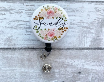 May include: A white retractable badge holder with a floral design and the name "Sandy" printed in black. The holder has a black retractable cord and a clear plastic clip.