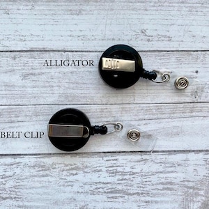 Op de afbeelding: Twee zwarte intrekbare badgehouders met alligatorclips en riemclips. De badgehouders bevinden zich op een wit houten oppervlak.