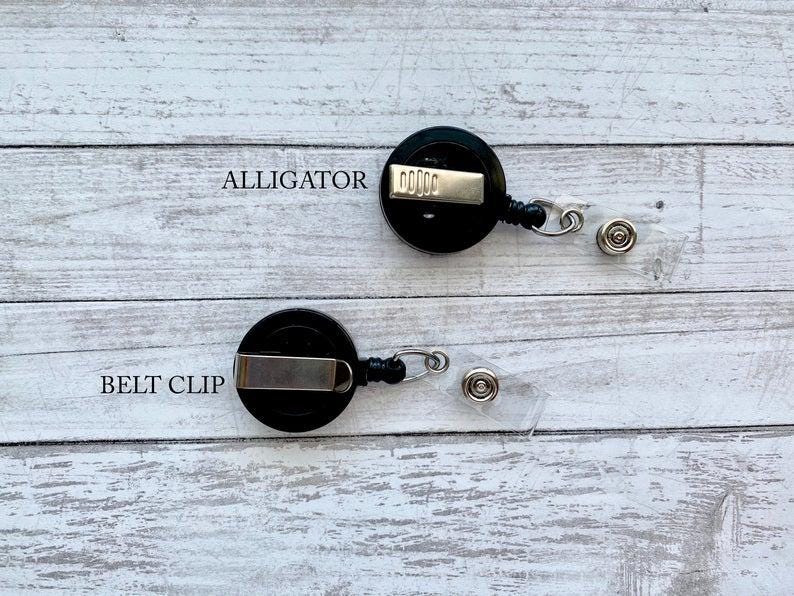 Op de afbeelding: Twee zwarte intrekbare badgehouders met alligatorclips en riemclips. De badgehouders bevinden zich op een wit houten oppervlak.