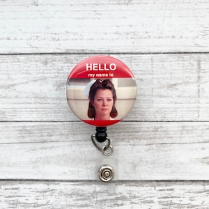 May include: A red and white button with the text "HELLO my name is" and a picture of a woman in a nurse's uniform. The button is attached to a black retractable badge holder.