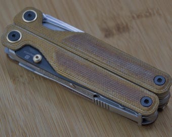 Könnte beinhalten: Ein Multitool mit einem strukturierten Griff in Beige und Braun sowie silbernen Werkzeugen. Das Werkzeug ist geschlossen und zeigt den Griff und die Drehgelenke. Der Griff hat ein abgenutztes Aussehen mit dunkelbraunen Akzenten.