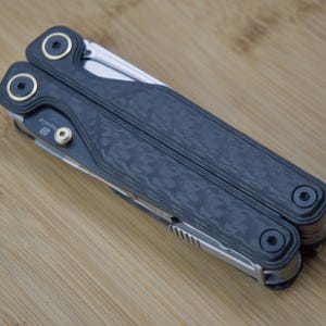 May include: A closed multi-tool with a dark grey textured handle and silver accents. The tool has gold-coloured circular accents and black screws. The multi-tool is lying on a light brown wooden surface.