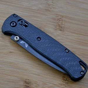 Könnte beinhalten: Ein Klappmesser mit einem dunkelgrauen Griff aus Kohlefaser und einer teilweise sichtbaren Metallklinge. Der Griff weist ein strukturiertes Webmuster, schwarze Schrauben, einen silbernen Daumenpin und eine Fangriemenöse am Ende auf.