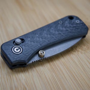 May include: A closed, dark grey pocket knife with a textured handle and a silver blade. The knife features a circular logo and a small button for opening and closing. The knife is lying on a wooden surface.