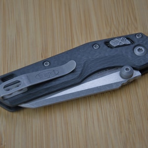 Peut inclure: Un couteau de poche pliant avec un manche en fibre de carbone sombre et un clip de poche en métal argenté. La lame est repliée dans le manche, révélant un bouton de pouce argenté et un mécanisme de verrouillage métallique sur le dessus.