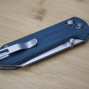 Könnte beinhalten: Ein geschlossenes Taschenmesser mit blauem Griff und silbernem Taschenclip. Die Klinge ist teilweise sichtbar, und das Messer liegt auf einer Holzoberfläche. Das Messer hat einen Knopf zum Öffnen und Schließen der Klinge.