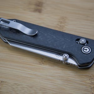 May include: A closed, dark grey folding knife with a silver pocket clip and a partially exposed blade. The knife has a textured handle and a silver button. The blade has a small logo.