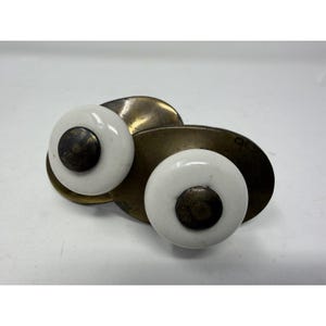 May include: Two cabinet knobs featuring round white ceramic centers with dark, weathered brass-toned metal caps in the middle. Each knob is mounted on an oval-shaped, tarnished brass-colored metal backplate, showcasing a vintage aesthetic.