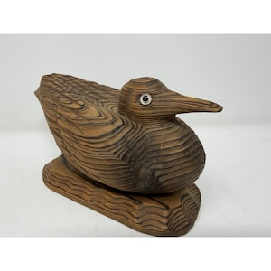 May include: A carved wooden bird figurine featuring a natural brown wood grain pattern with visible rings. The bird has a pointed beak, a rounded body, and a single inset eye with a dark center and a metallic silver rim, mounted on a base.