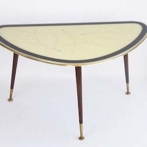 Könnte beinhalten: Ein halbmondförmiger Couchtisch im Mid-Century-Modern-Stil. Der Tisch hat eine cremefarbene Platte mit schwarzem Rand und Messingverzierungen. Die Beine sind aus dunklem Holz mit messingfarbenen Spitzen. Der Tisch ist ca. 76 cm breit.