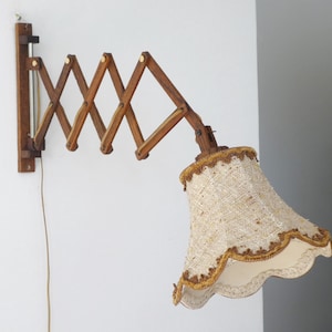 May include: A wall-mounted lamp featuring a brown wooden accordion-style scissor arm that extends outwards. The lamp includes a textured, cream-coloured fabric lampshade with a scalloped edge and decorative brown trim, connected to a thin power cord.
