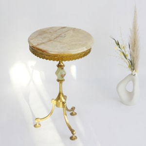 May include: A small, antique side table with a round, marble top. The table has a gold-coloured, ornate metal base with three legs. The table's top is approximately 30 cm in diameter. A white vase with dried flowers is to the right.