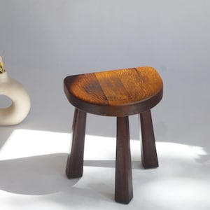 Puede incluir: Un taburete de madera pequeño de tres patas con un asiento semicircular que presenta un acabado de veta de madera marrón oscuro. El taburete tiene tres patas cónicas y una superficie superior lisa y redondeada que muestra la textura natural.