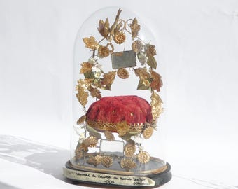 May include: A decorative display piece under a glass dome. It features a red velvet cushion, surrounded by gold-coloured floral and leaf embellishments. The base is black with a label that includes text and the year 1936.