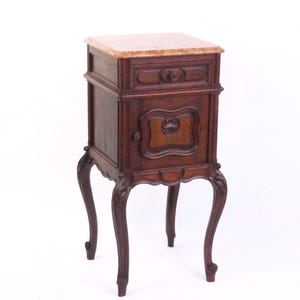 Peut inclure: Une table de chevet en bois brun foncé dotée d'un plateau en marbre clair, d'un tiroir rectangulaire avec un bouton central et d'une porte de placard avec une poignée décorative. Le meuble repose sur quatre pieds galbés.