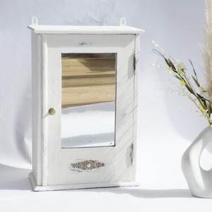 May include: A white, distressed-finish wall-mounted cabinet with a mirrored door. The cabinet has a small brass knob and decorative detailing at the bottom. A white, abstract vase holds dried flowers and pampas grass.