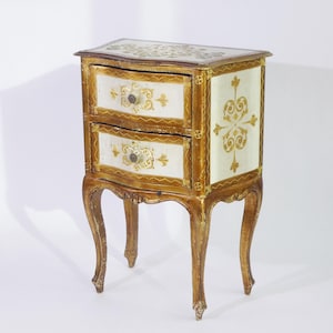Op de afbeelding: Een kleine, antieke nachtkast met twee lades. De tafel is crème en goud geschilderd, met sierlijke gouden details op de lades en zijkanten. De poten zijn gebogen en de bovenkant is versierd met een gouden bloemmotief. Ongeveer 60 cm hoog.