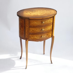 May include: An antique wooden side table with three drawers. The table has a light brown finish with floral inlay details. The legs are curved and the drawer pulls are brass. The table is approximately 76 cm tall.