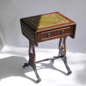 May include: A dark wood side table featuring a rectangular top with a green leather inlay, a single drawer with two circular metal pulls, drop-leaf sides, and four curved legs ending in claw-and-ball feet with small castors for mobility.