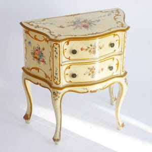 May include: A small, cream-colored chest of drawers with two drawers and curved legs. The chest is adorned with hand-painted floral designs in various colors and gold trim. The hardware is dark brown.