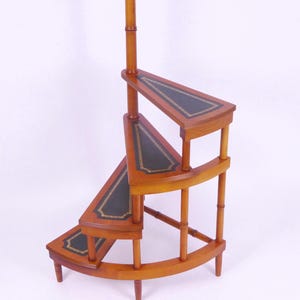 Puede incluir: Una estantería de esquina de madera con tres niveles. Cada nivel está forrado con cuero negro y tiene un ribete dorado decorativo. La estantería está hecha de madera clara y tiene un acabado natural.