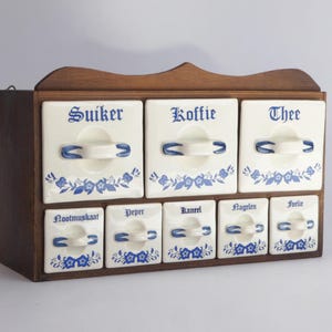 Peut inclure: Un ancien pot à épices avec huit tiroirs en céramique blanche. Chaque tiroir est étiqueté avec un mot néerlandais en script bleu et présente un motif floral bleu. Le support est logé dans un cadre en bois brun foncé.