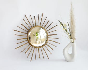 May include: A gold-coloured sunburst mirror with a round, reflective centre. The mirror is mounted on a white wall, next to a white vase holding dried flowers and pampas grass. The mirror's rays extend outwards.