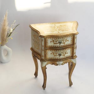 May include: A small wooden bedside table featuring two drawers with gold and cream painted floral scrollwork. The piece stands on four curved legs and includes small metal drawer pulls. A white ceramic vase with dried pampas grass is nearby.
