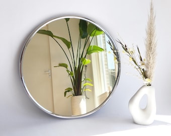 May include: A round mirror with a polished silver frame reflects a room with a large potted plant and a window. A white vase with a unique opening holds dried flowers and pampas grass, adding a touch of natural decor.