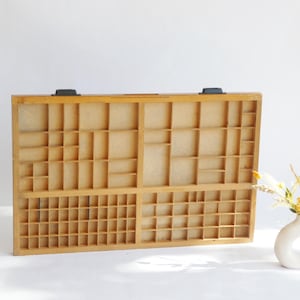 May include: A rectangular wooden printer's tray featuring numerous small, varied compartments arranged in a grid pattern. The light brown wood has a natural finish, and two black metal hardware pieces are attached to the top edge of the frame.