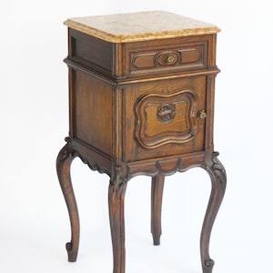 May include: A dark wood bedside table with a marble top, a single drawer, and a cabinet door with a carved design. The table has four ornate legs.