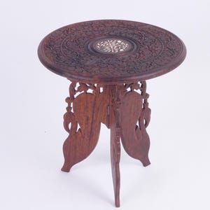 May include: A dark brown, round, wooden side table with intricate floral carvings on the tabletop. The table has three ornate legs and a decorative center inlay. The table is approximately 61 cm in diameter and 71 cm tall.