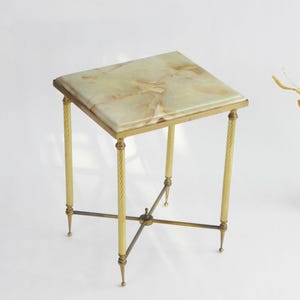 Puede incluir: Una mesa auxiliar cuadrada con tapa de mármol y patas de metal dorado. La parte superior de la mesa es de color verde claro y beige. Las patas tienen un diseño de cuerda trenzada y están conectadas por una barra transversal en la parte inferior. La mesa mide unos 51 cm de alto.