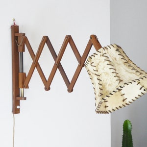 Op de afbeelding: Een houten wandlamp met een opvouwbare arm en een crèmekleurige stoffen kap met bruine stiksels.