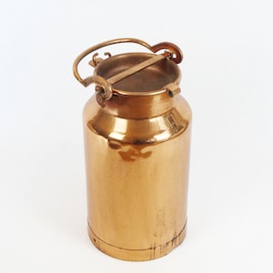 Puede incluir: Una pequeña lata de leche de metal de color dorado con una tapa con bisagras y una asa de transporte. La lata tiene una superficie lisa y brillante.