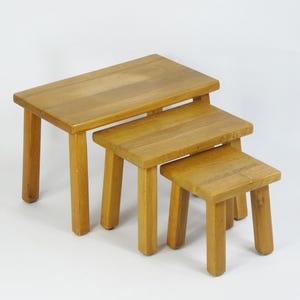 Puede incluir: Un conjunto de tres mesas nido de madera. Las mesas son de madera marrón claro y tienen un diseño simple y rectangular. La mesa más grande está en la parte inferior y la mesa más pequeña está en la parte superior.