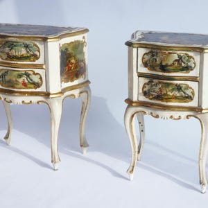 May include: A pair of antique white bedside cabinets, each with two drawers. The drawers and sides are decorated with painted landscape scenes and gold trim. The cabinets have curved legs and gold trim.