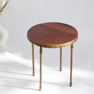 May include: A round side table with a dark brown wood grain top and a gold-toned metal frame. The table features a decorative, ornate scalloped edge along the perimeter of the tabletop and four slender, straight metal legs with small feet.