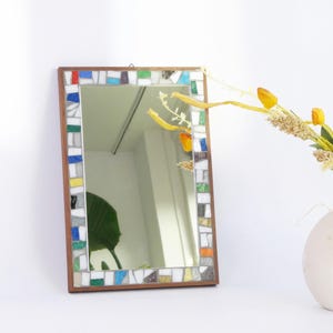 May include: Rectangular mirror with a mosaic tile border in shades of red, blue, green, yellow, and gray. The mirror has a wooden frame and is hanging on a white wall. A vase with yellow flowers is in the background.